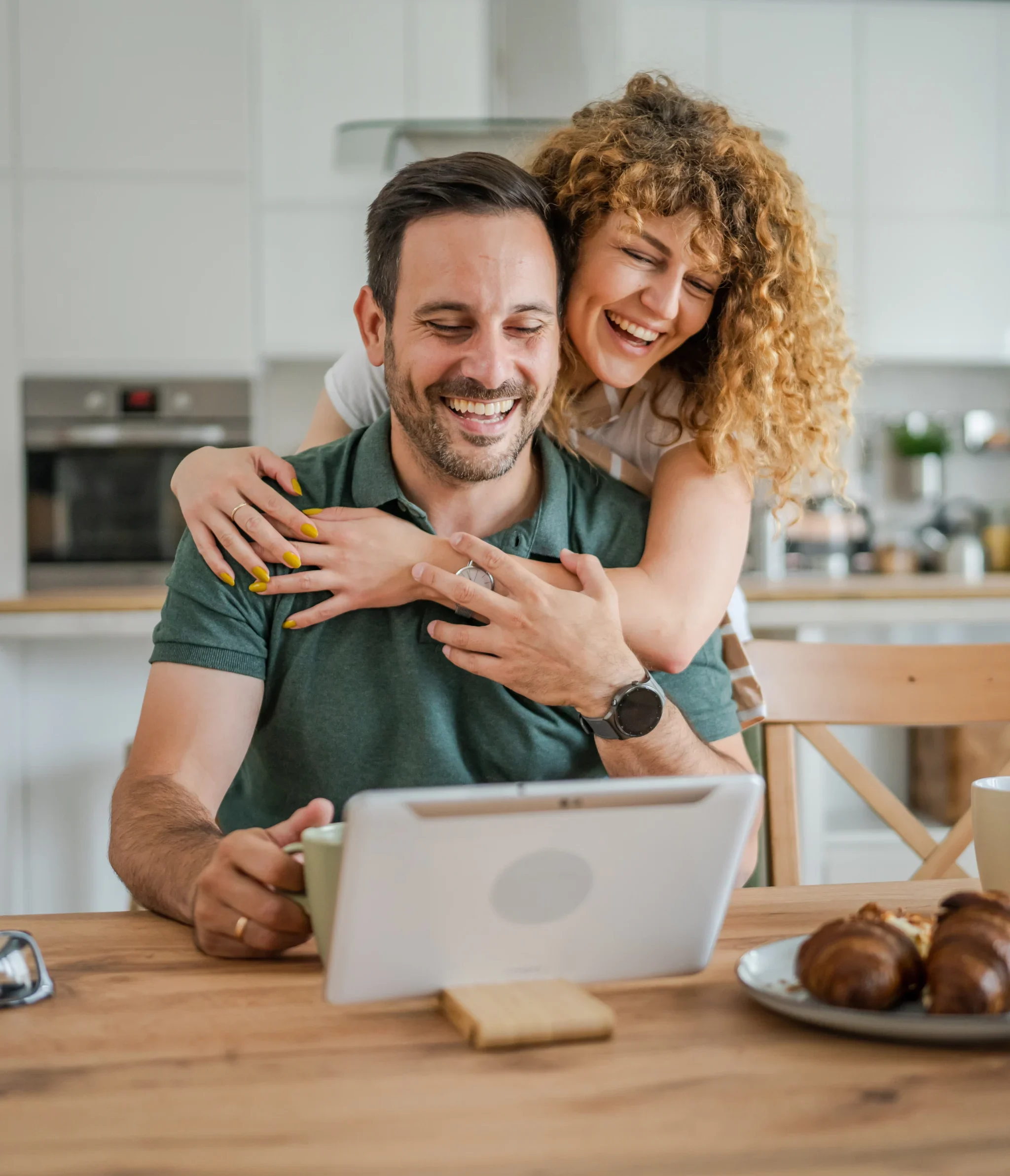 Happy couple laughing together at home while looking at a tablet during breakfast.