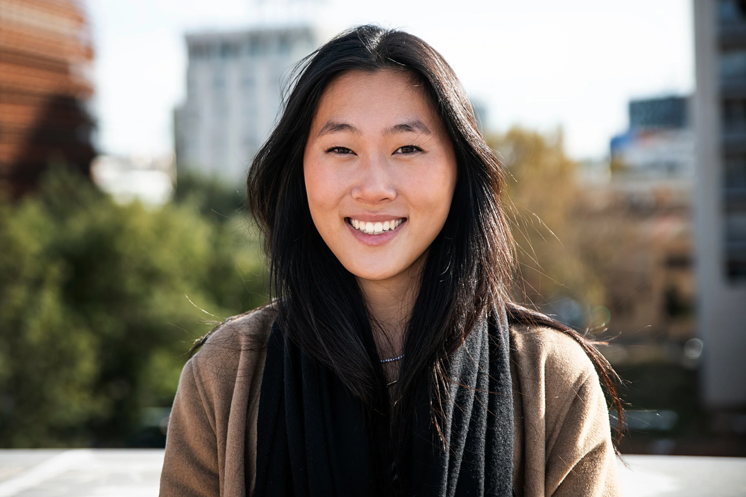Smiling young woman outdoors with city skyline in the background on a sunny day.