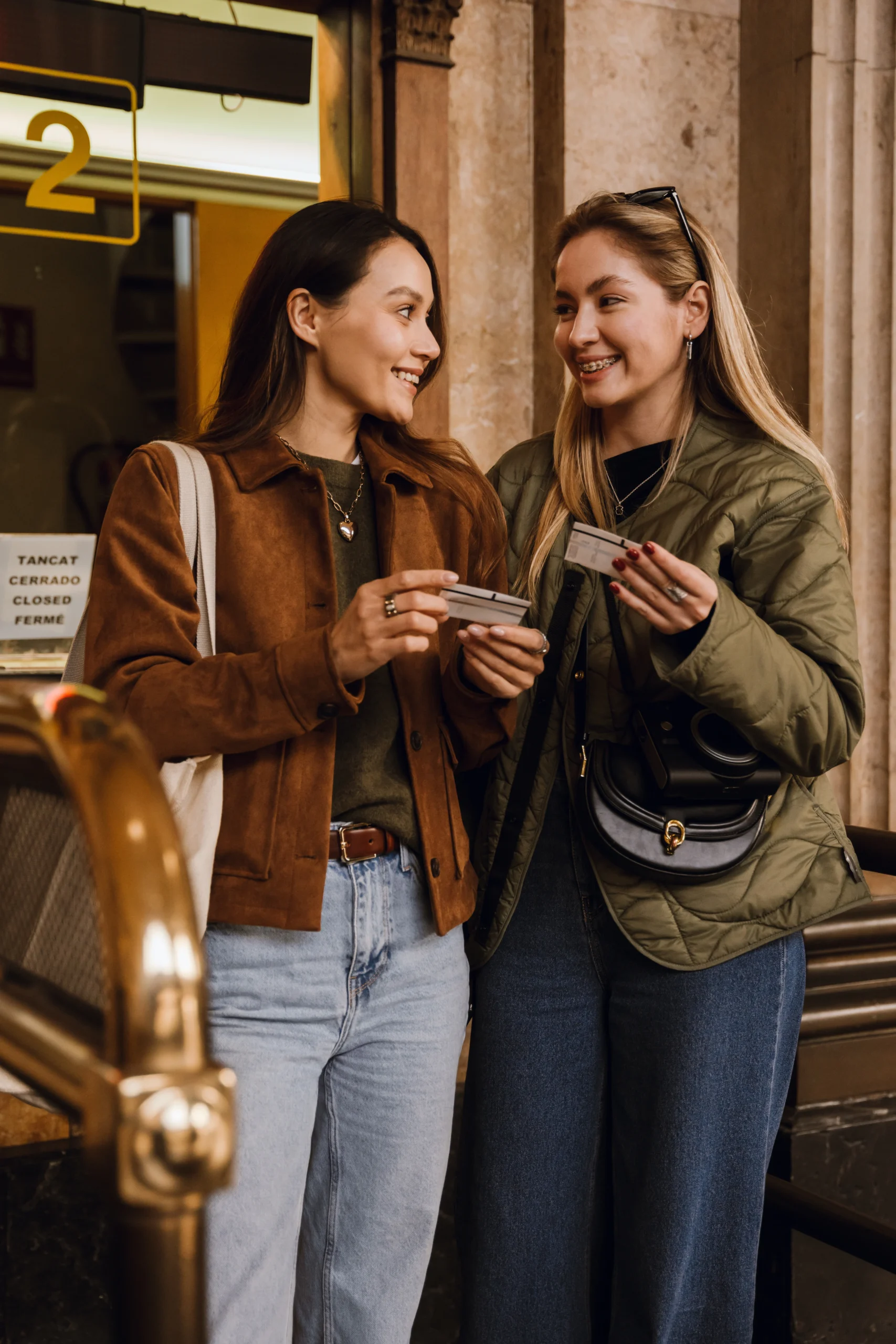 Two young women smiling and chatting while holding tickets outside a building.