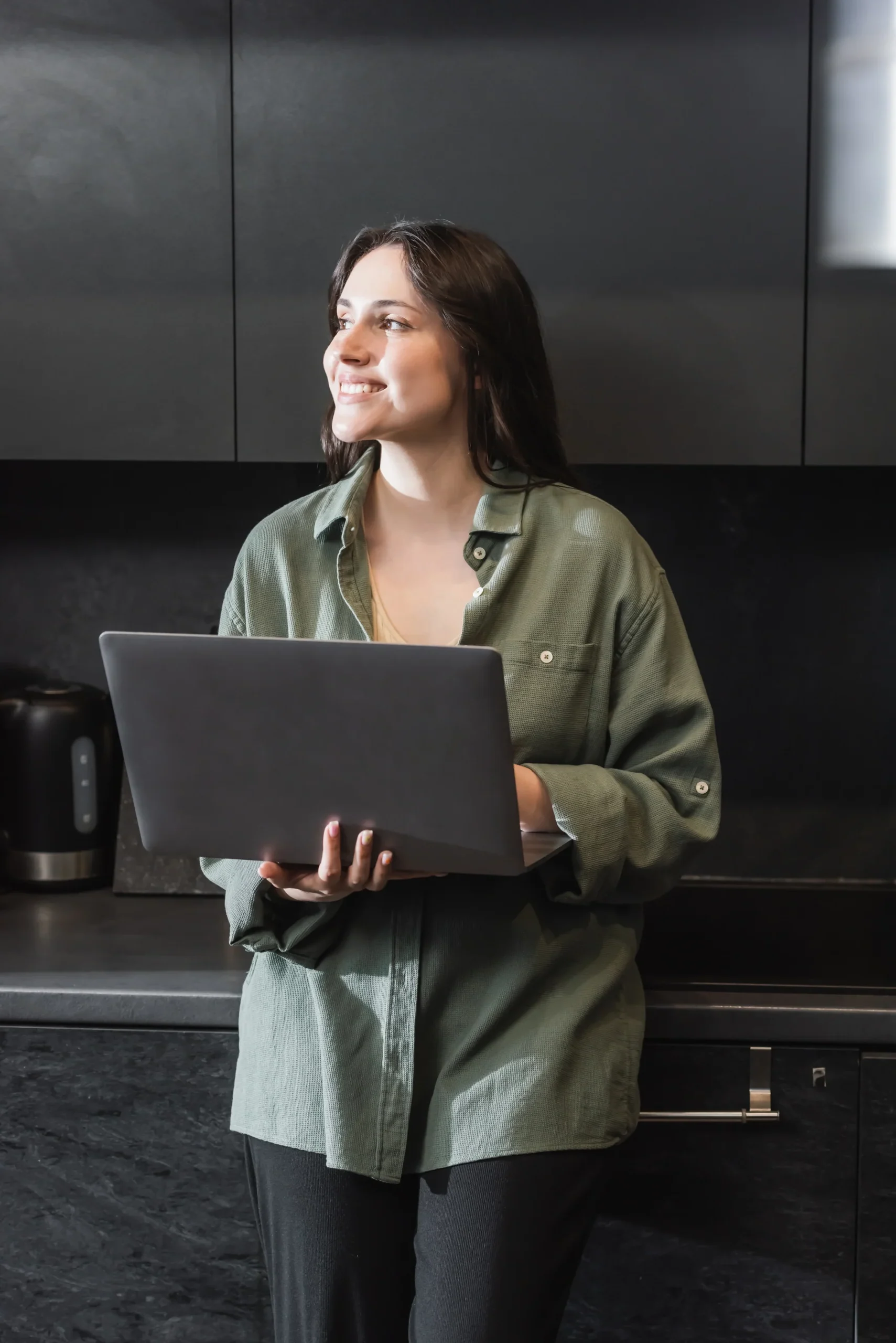 Young woman smiling while holding a laptop in a modern kitchen.