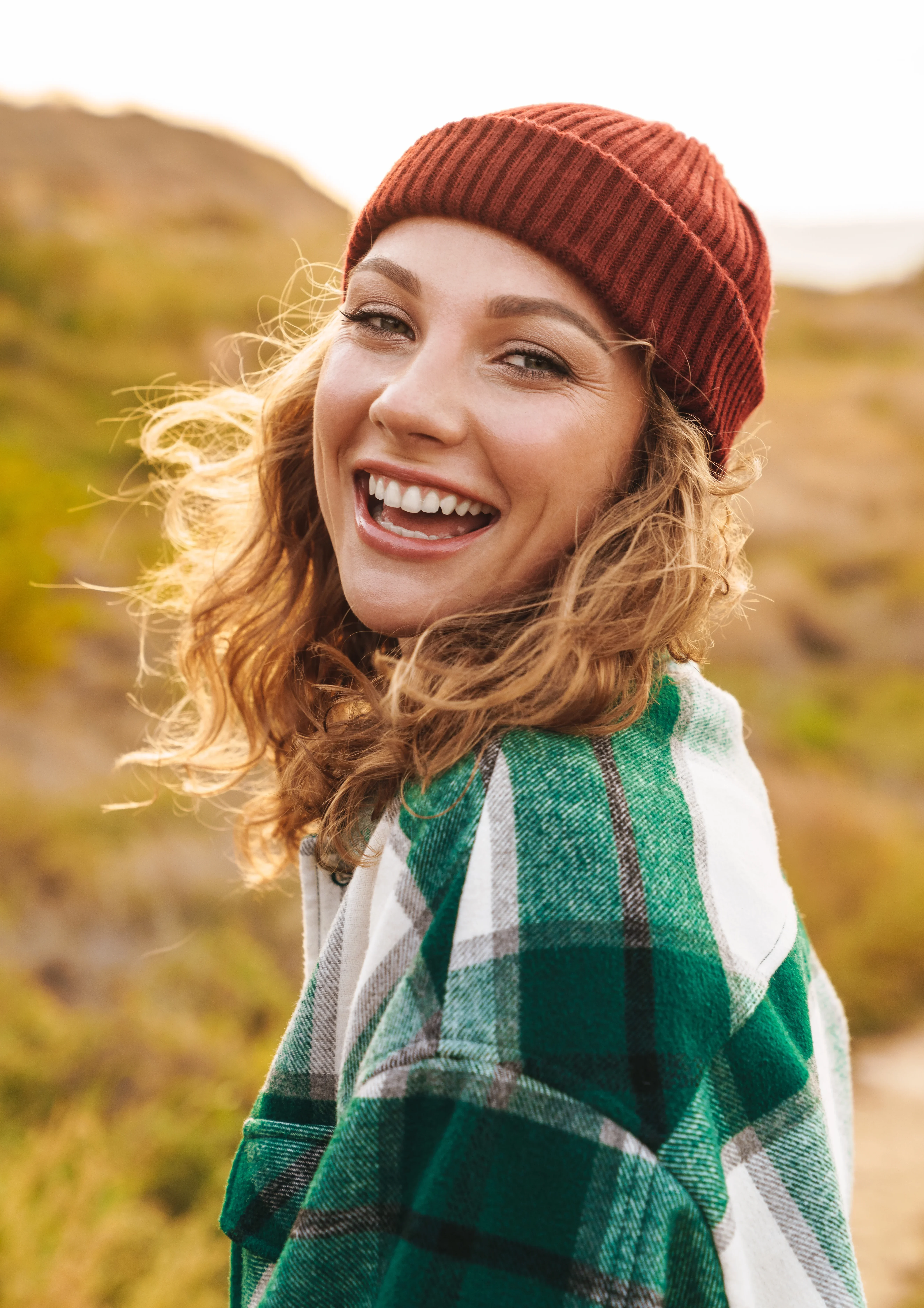 Smiling young woman wearing a red beanie and green plaid jacket outdoors.