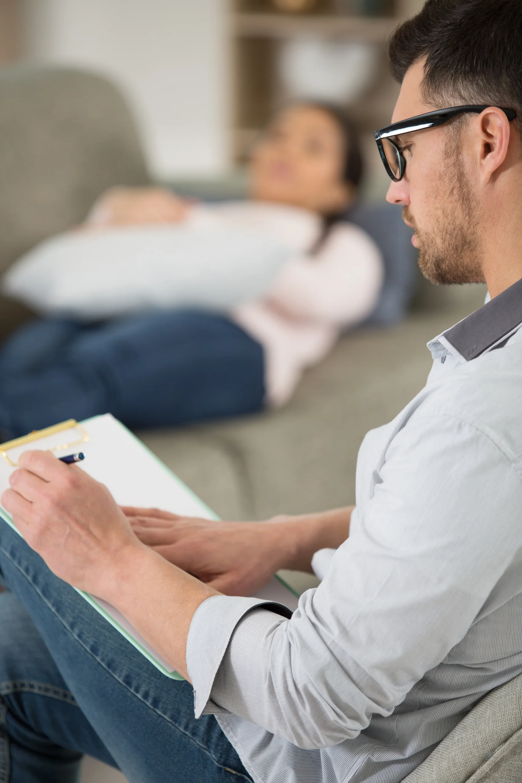 Psychiatrist taking notes during a one-on-one therapy session with a patient resting on a couch.