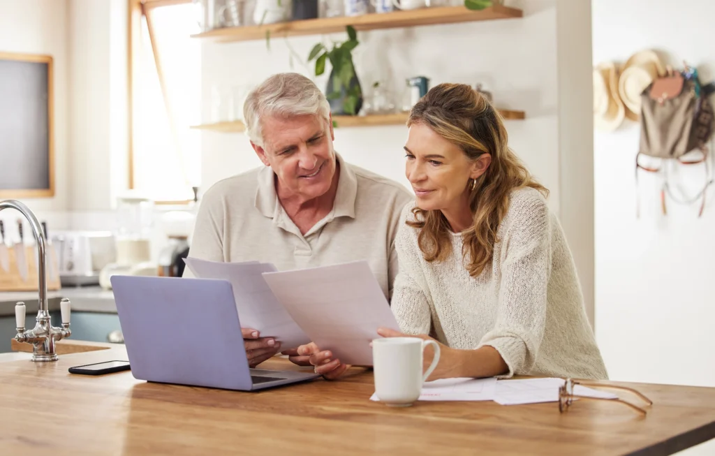 Couple reviewing bills and paperwork at a kitchen table with coffee and a laptop