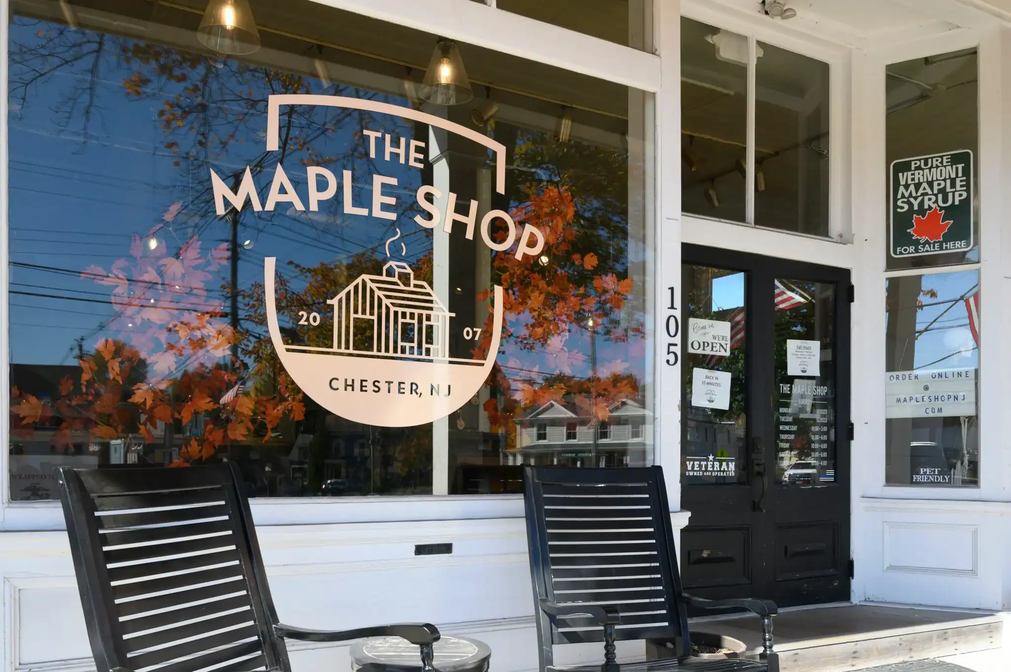 Front entrance of The Maple Shop in Chester, New Jersey, with porch chairs and fall foliage reflected in the storefront window.