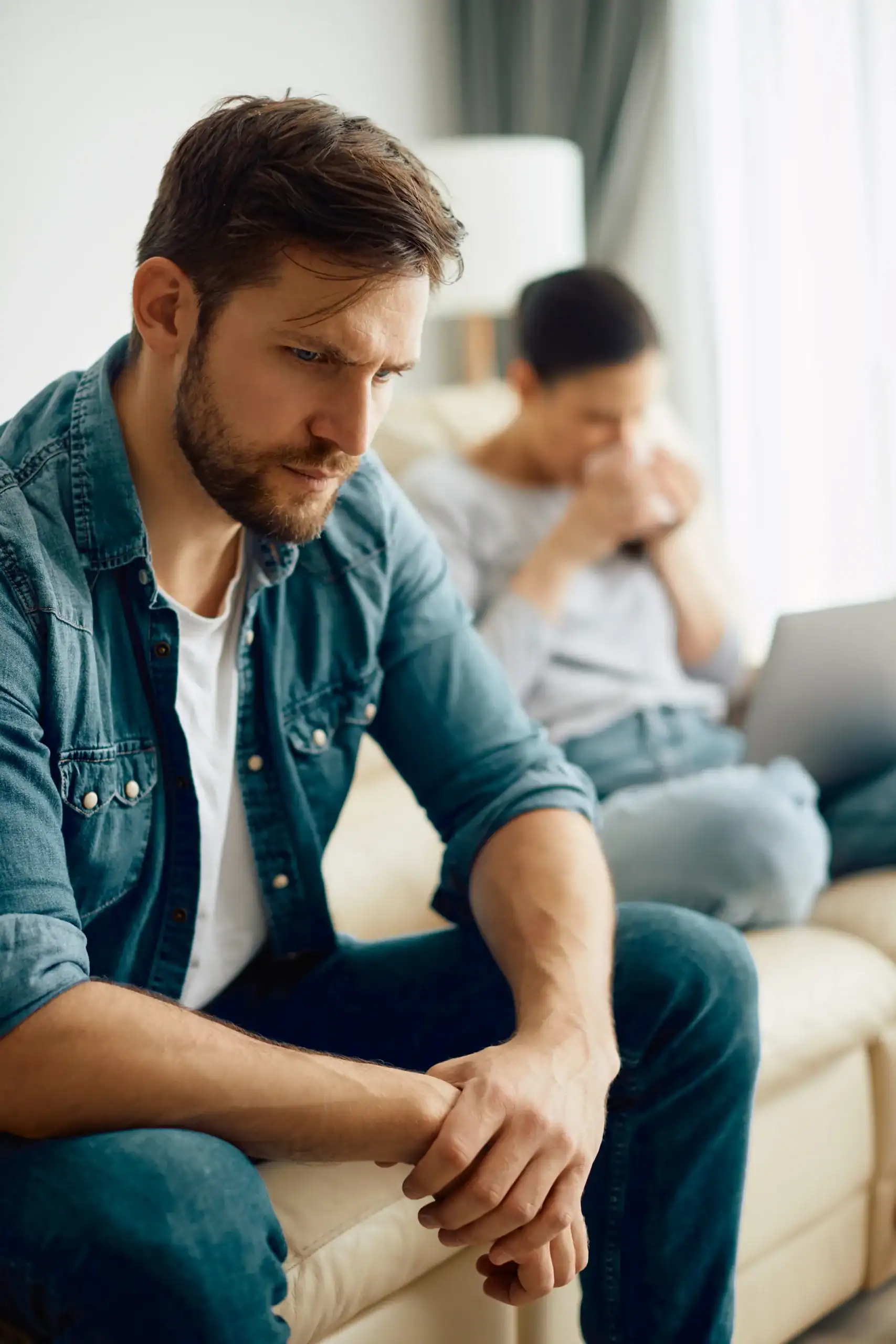 Man sitting on a couch looking distressed while partner is upset in the background