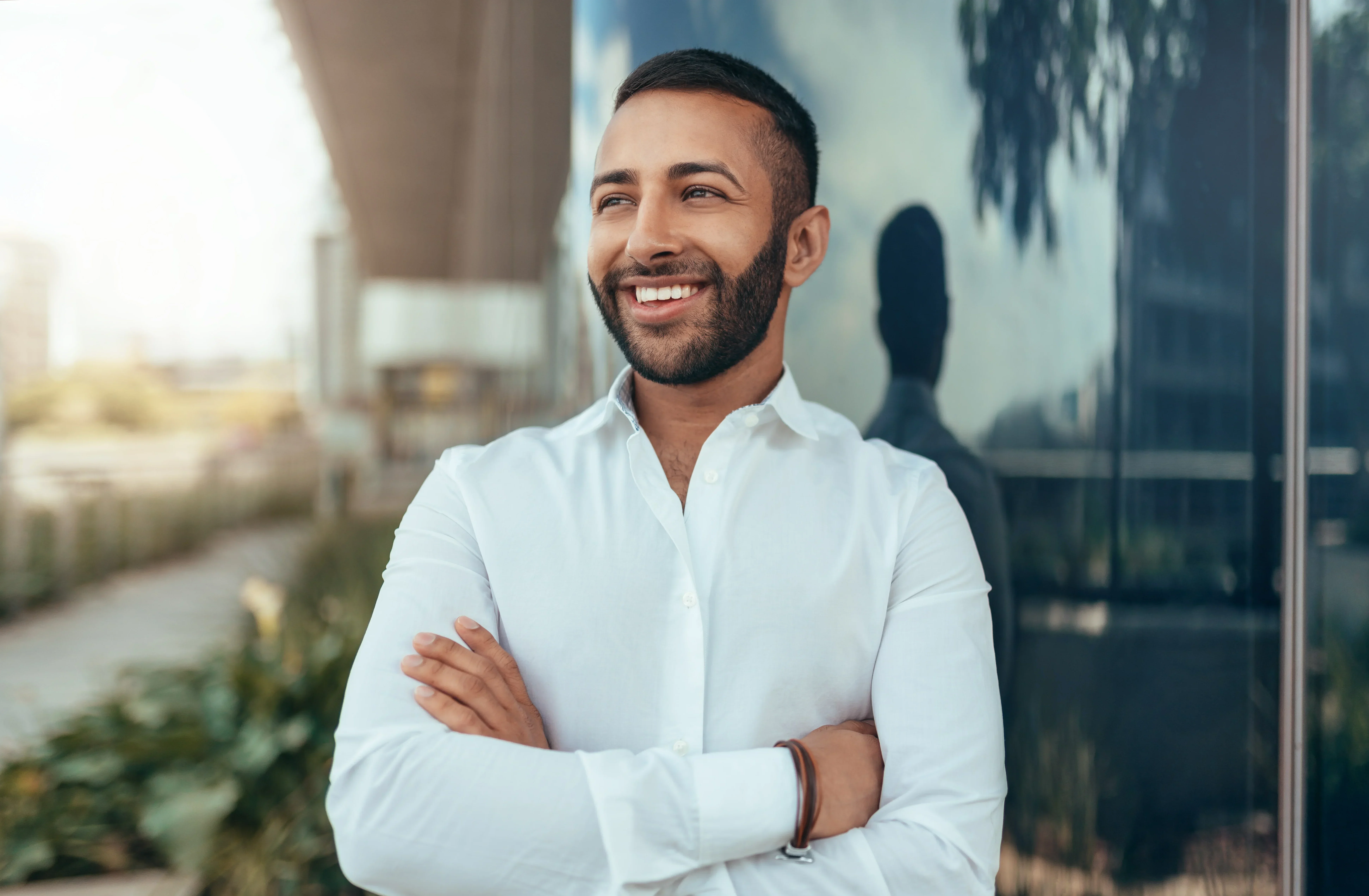 Smiling man in a white shirt with arms crossed standing outdoors near a modern building