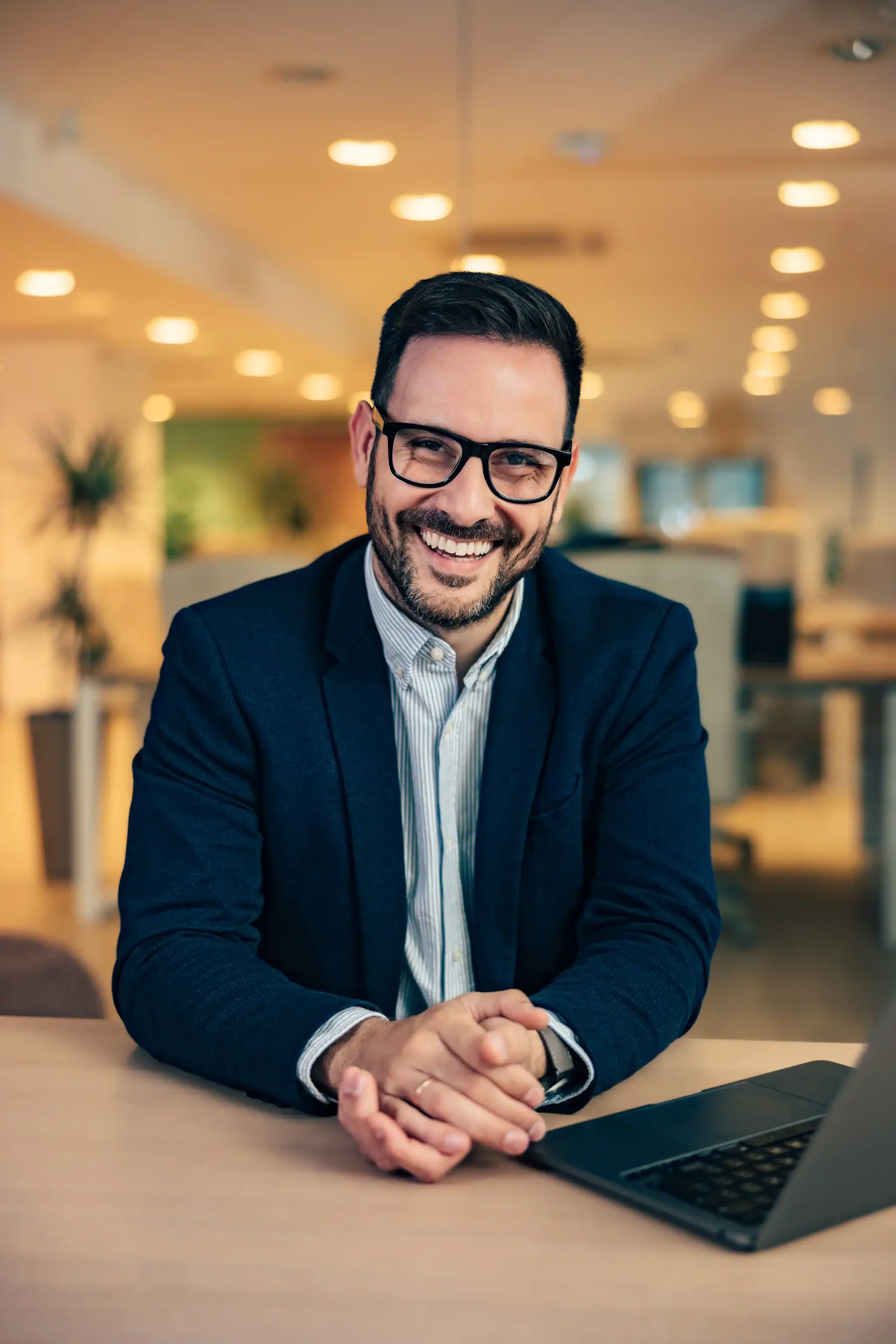 Smiling professional man wearing glasses and a suit, sitting at a desk with a laptop in a modern office.