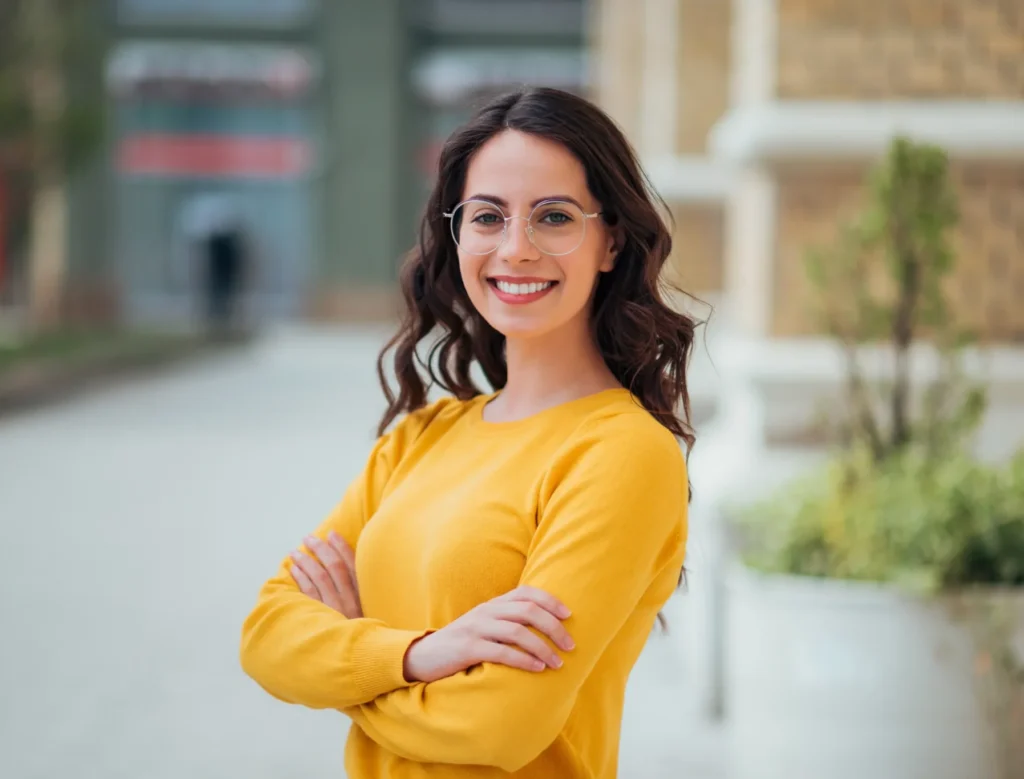 Smiling woman wearing glasses and a yellow sweater with arms crossed outdoors