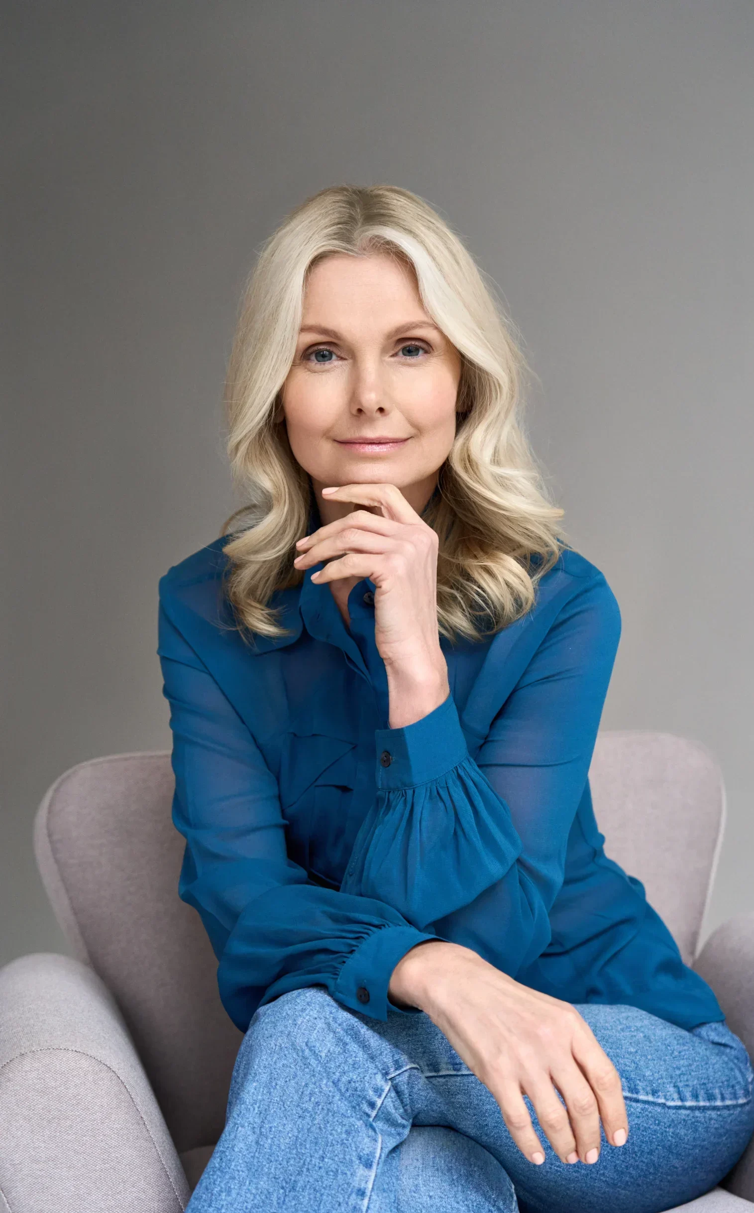 Blonde woman in a blue blouse sitting in an armchair with hand on chin