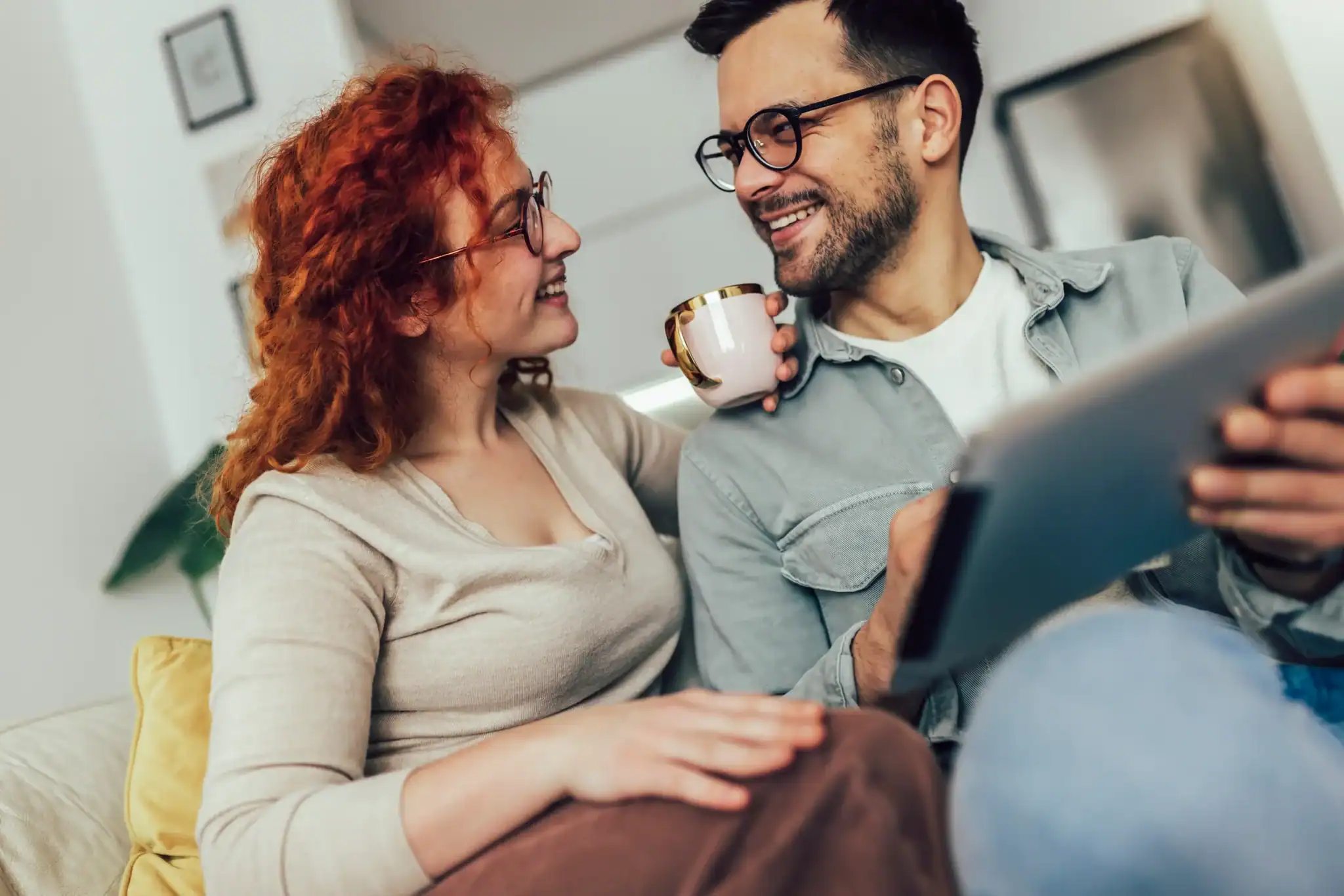 Couple sitting on a couch smiling together while holding a tablet and a coffee mug
