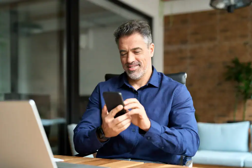 Man sitting at a desk smiling while using a smartphone next to a laptop