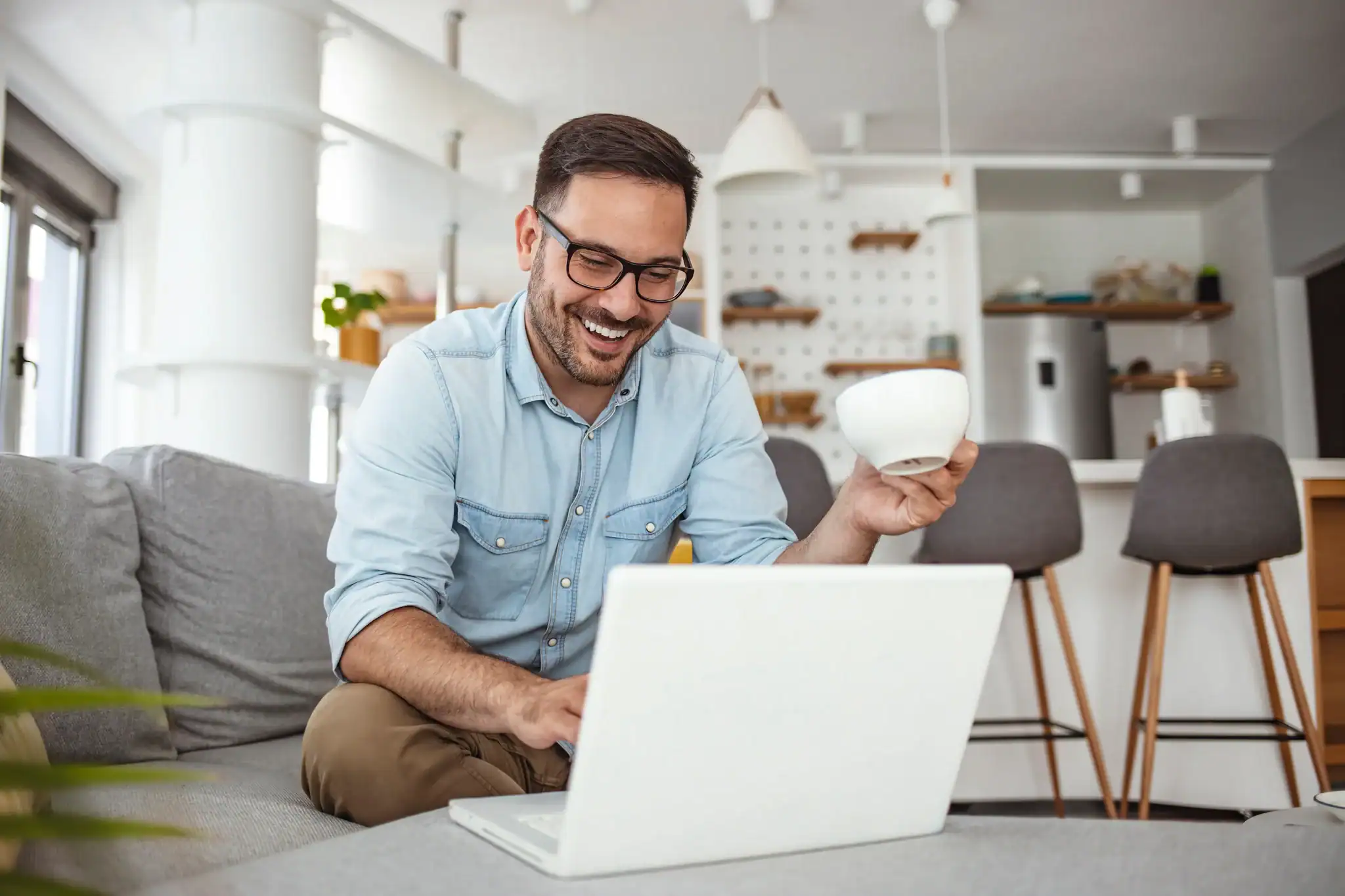 Smiling man sitting on a couch using a laptop while holding a coffee cup at home