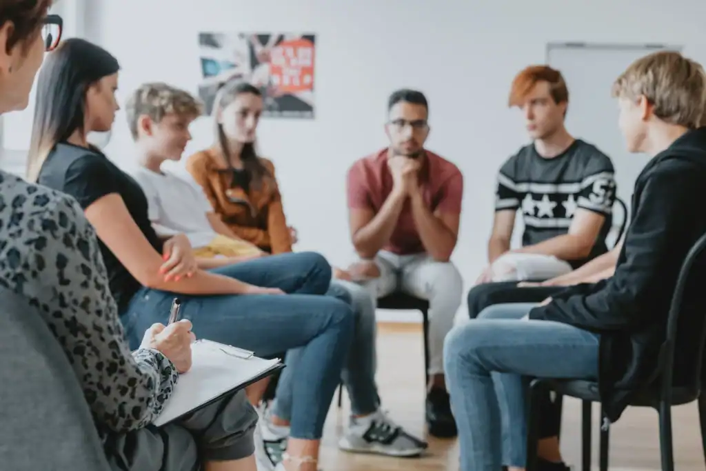Clinician with clipboard leading a structured group therapy session while participants listen in a circle