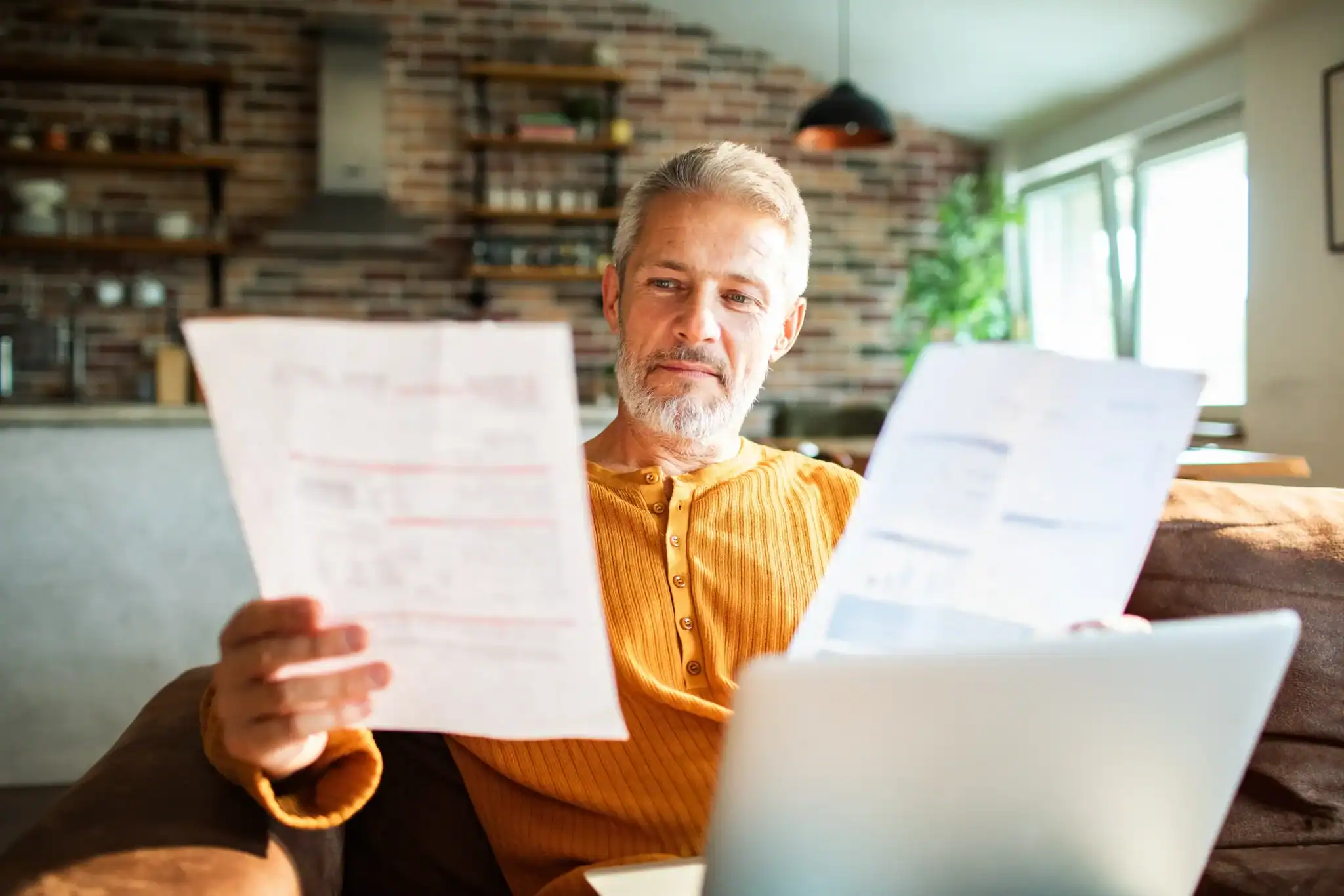 Man reviewing insurance paperwork and medical bills on his couch while researching depression treatment costs on a laptop