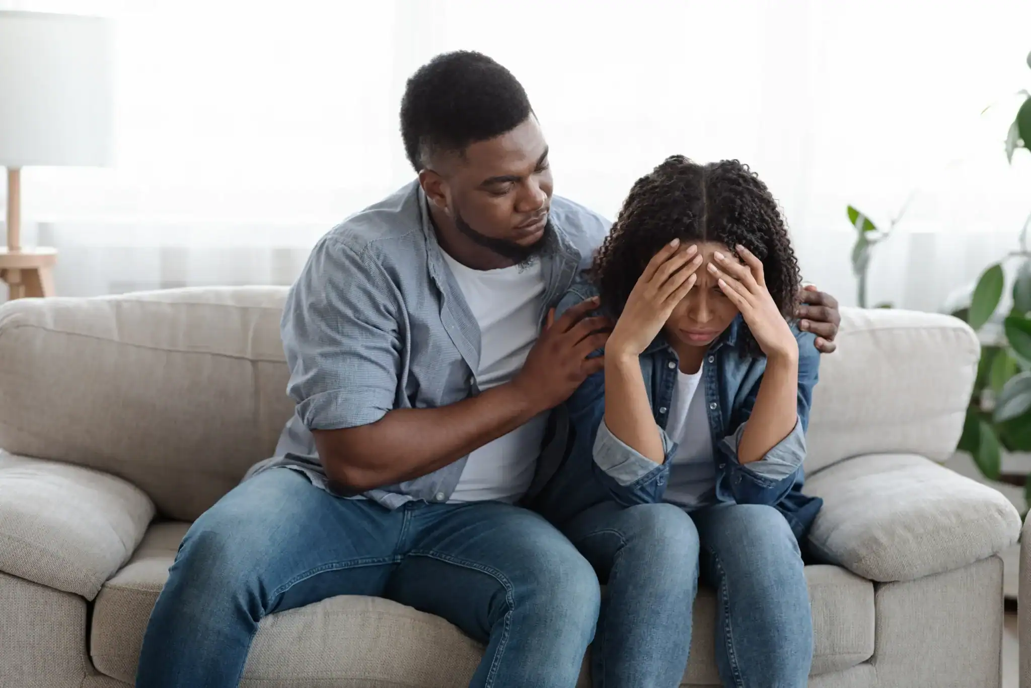 A man sits beside his distressed partner on a couch at home, placing a supportive hand on her shoulder as she holds her head in her hands.