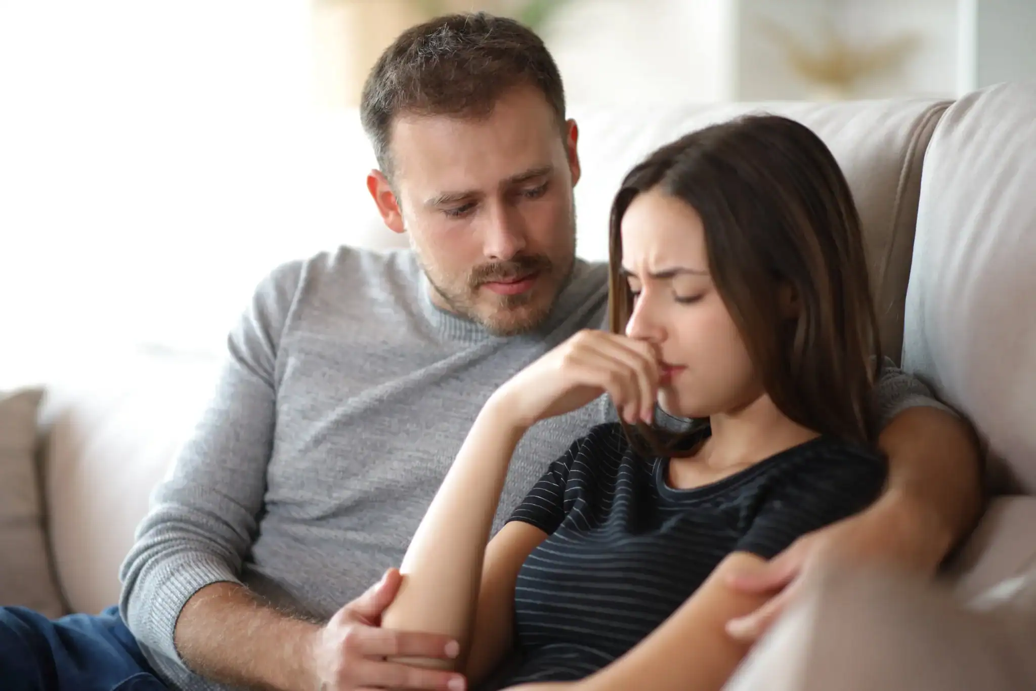 Partner sitting beside an anxious spouse on a couch, gently holding her arm in a quiet moment of supportive presence. Partner sitting beside an anxious spouse on a couch, gently holding her arm in a quiet moment of supportive presence.