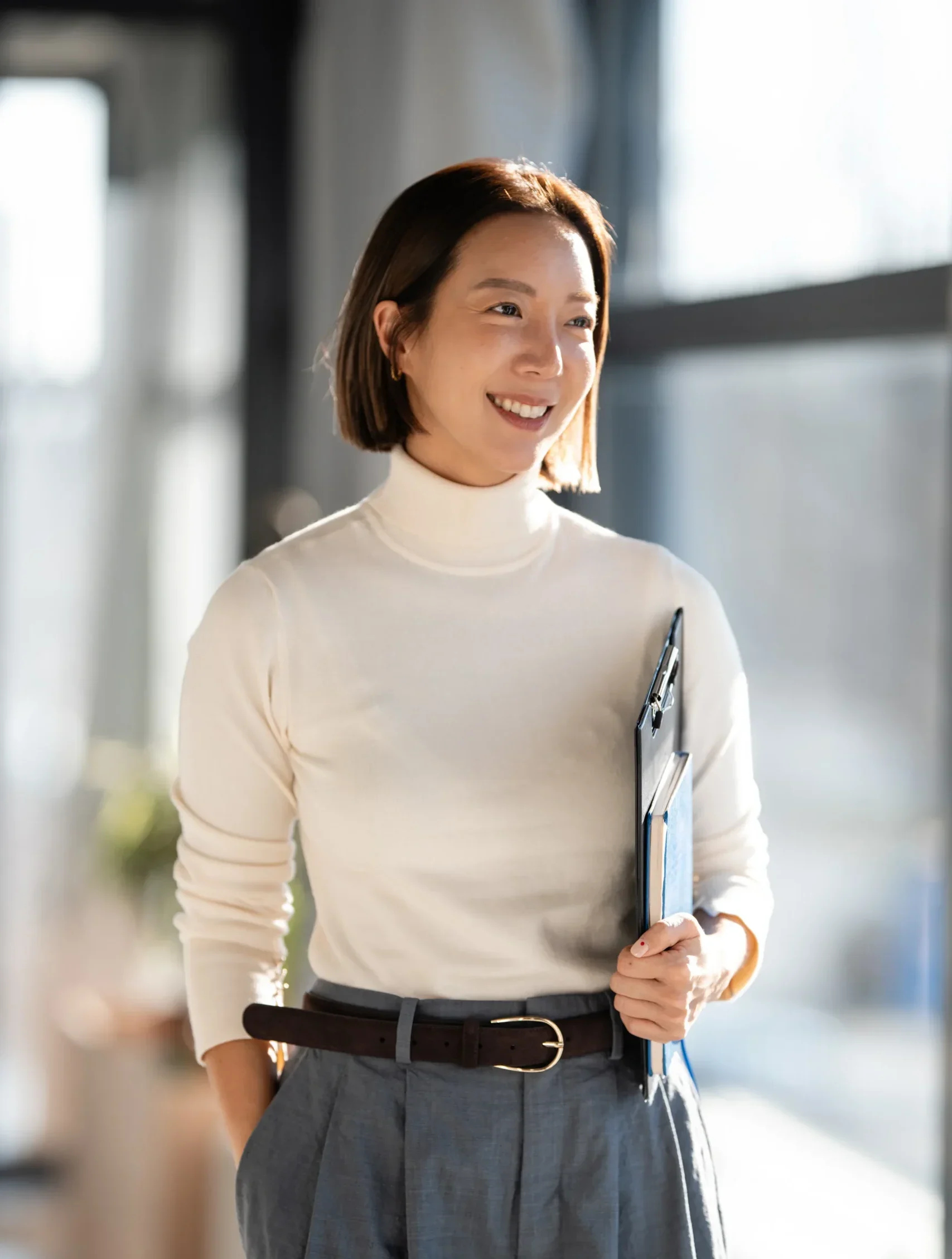 A professional holding a clipboard and notebook smiles while standing in a sunlit office.