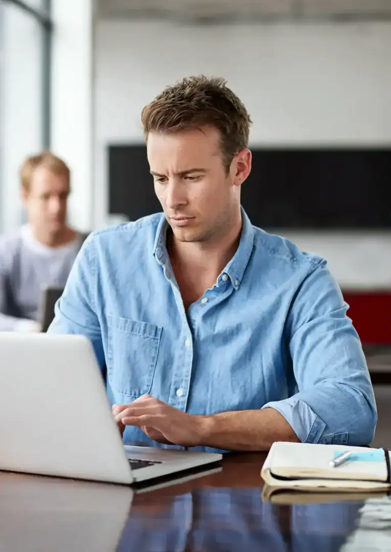 A man focused on a laptop at a table, with a notebook and pen beside him.