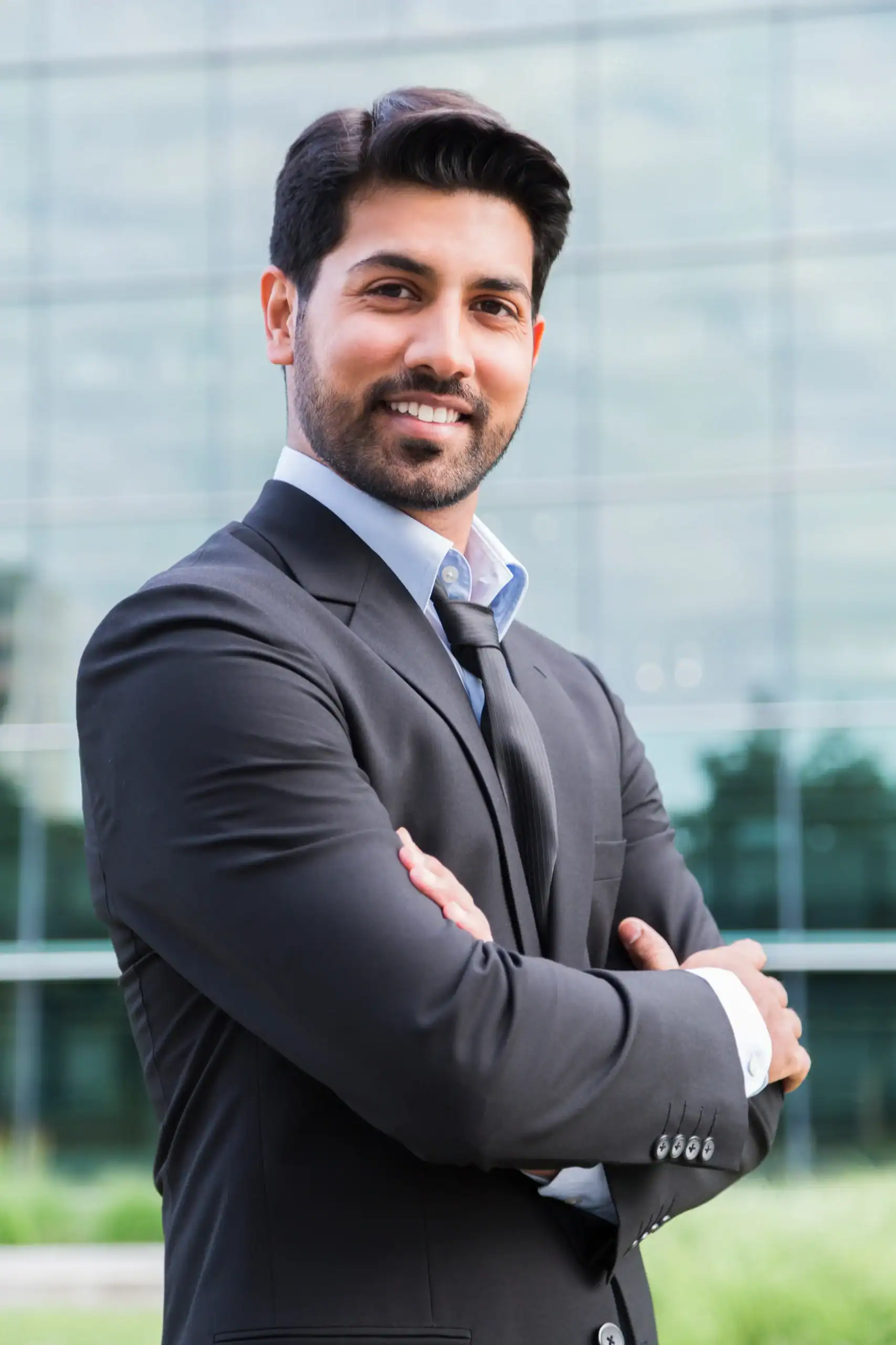 A man in a dark suit smiles with his arms crossed outside a glass office building.