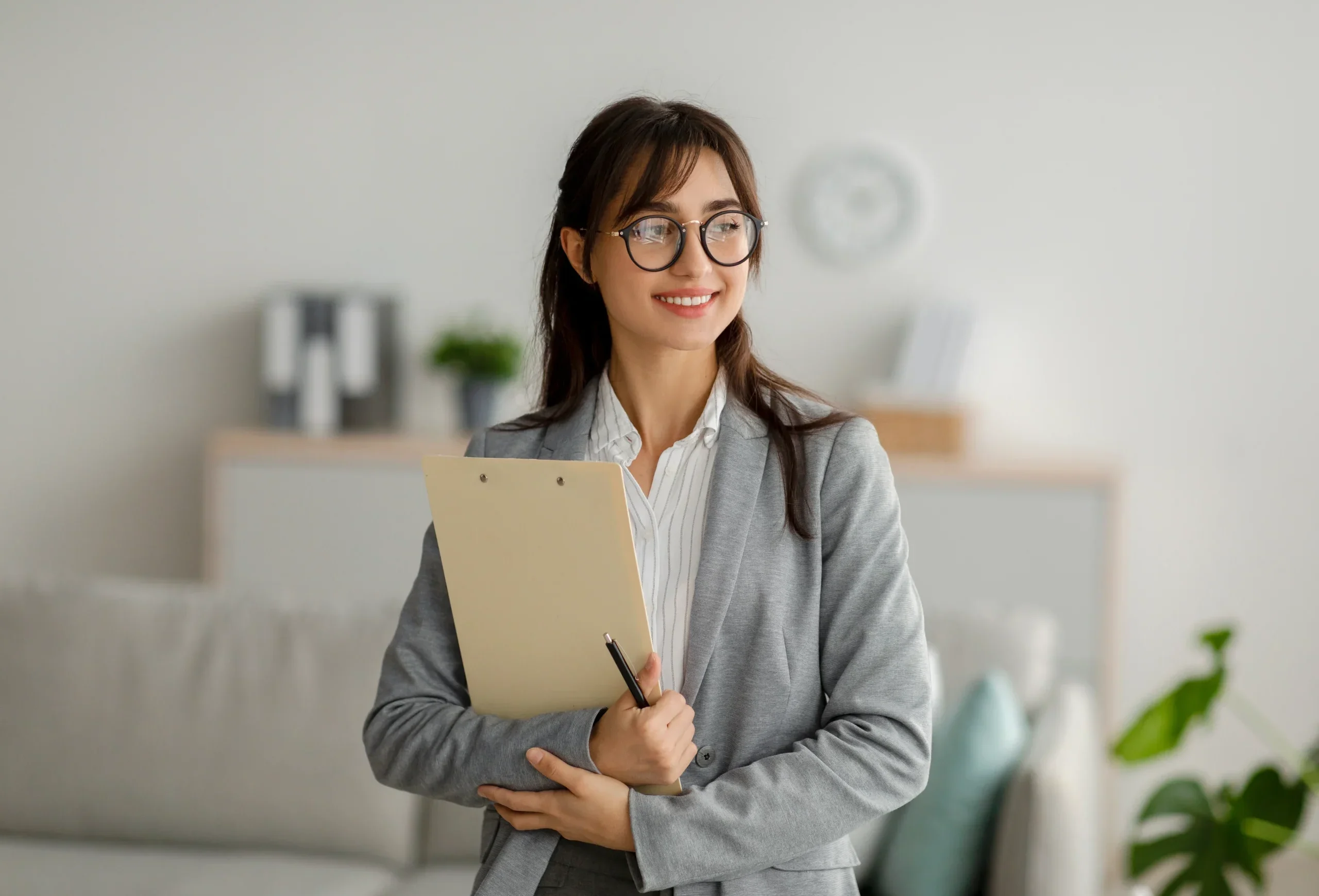 Smiling mental health professional in a grey blazer holding a clipboard and pen.