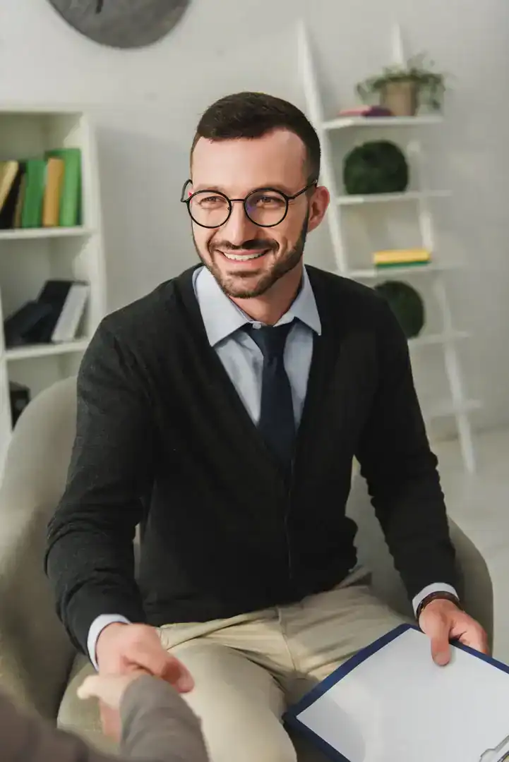 A smiling man with a clipboard shakes hands with a person while seated in a bright office.