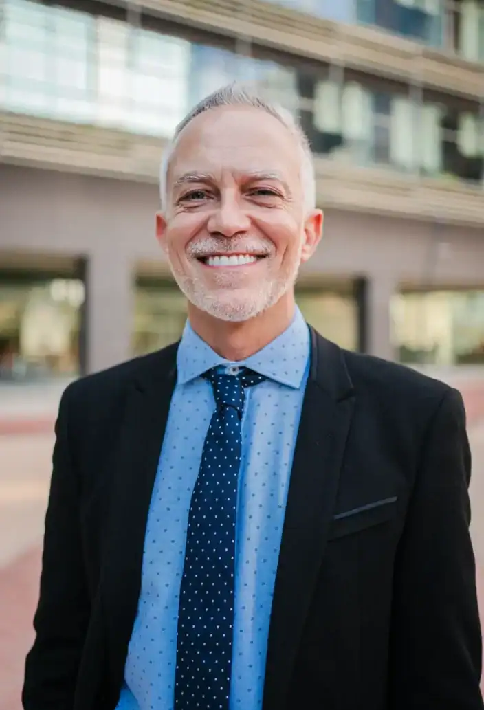 A smiling man in a dark blazer and tie standing outdoors in front of a modern office building.