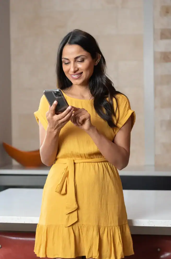 A smiling woman using her smartphone, with warm lights blurred behind her.