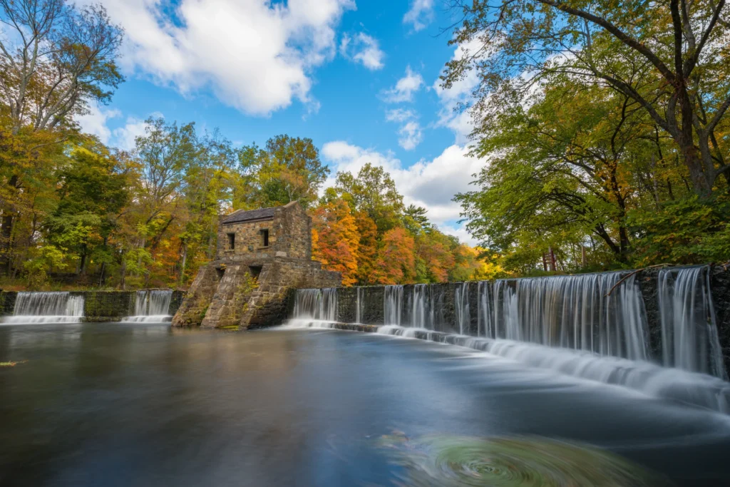 Speedwell Dam in Morristown, NJ, with water flowing over the falls and autumn foliage along the shoreline.