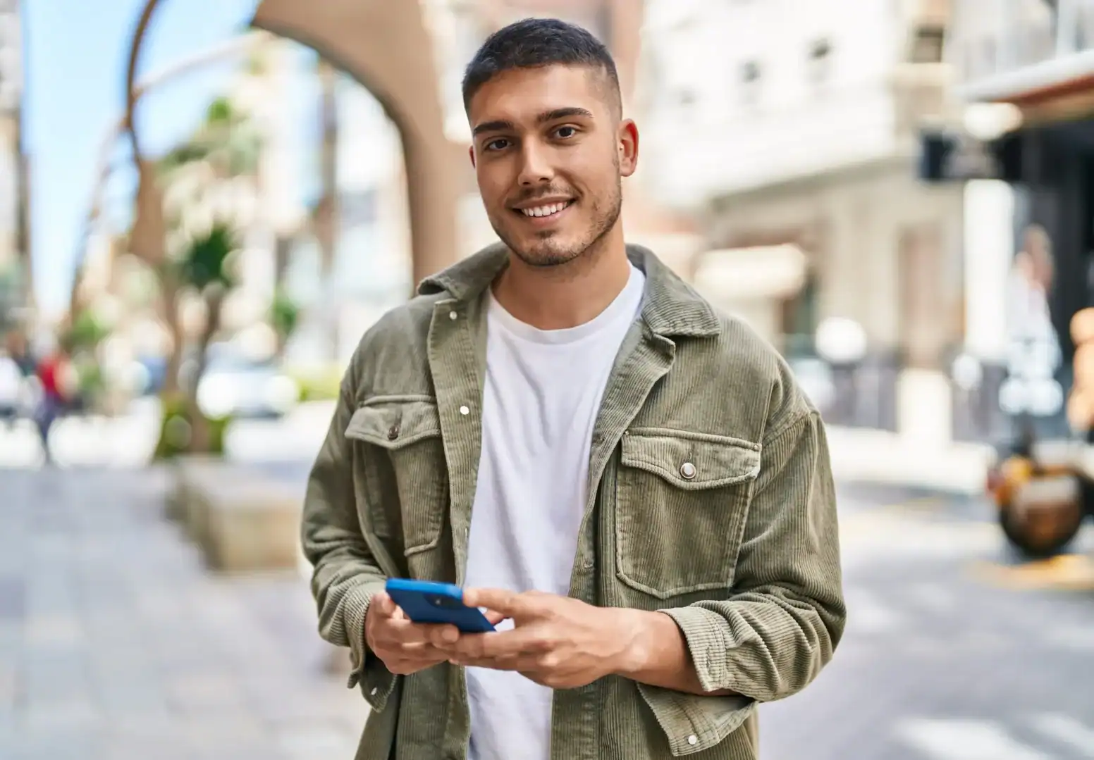 Smiling young man standing on a sidewalk holding a smartphone.