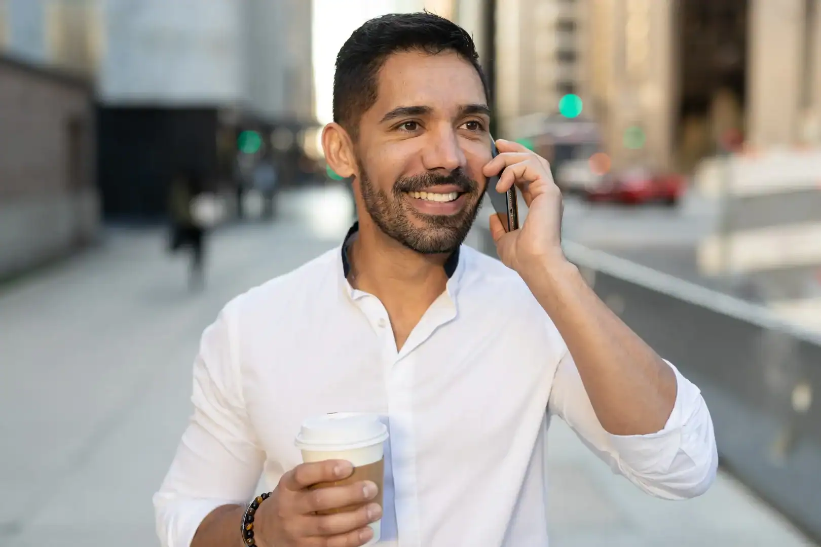 Smiling man talking on the phone outdoors while holding a coffee.