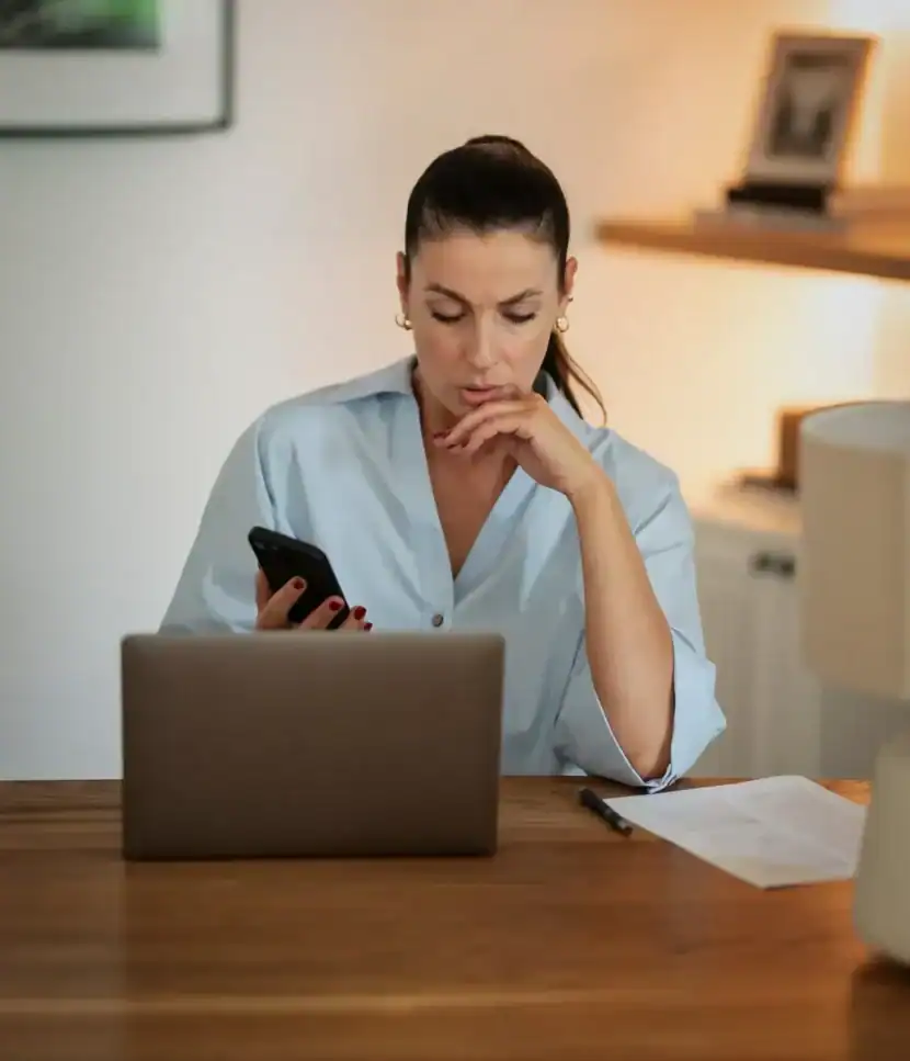 Woman at a desk checking her phone while reviewing a document next to her laptop.