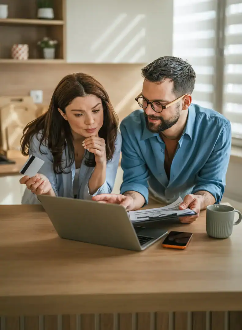 A couple at a kitchen table review paperwork on a laptop, one holding a card and the other pointing at a form.