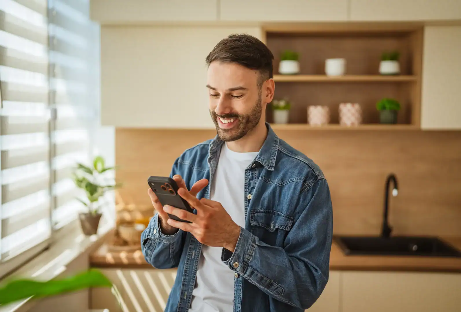 A smiling man stands in his kitchen looking at his phone.