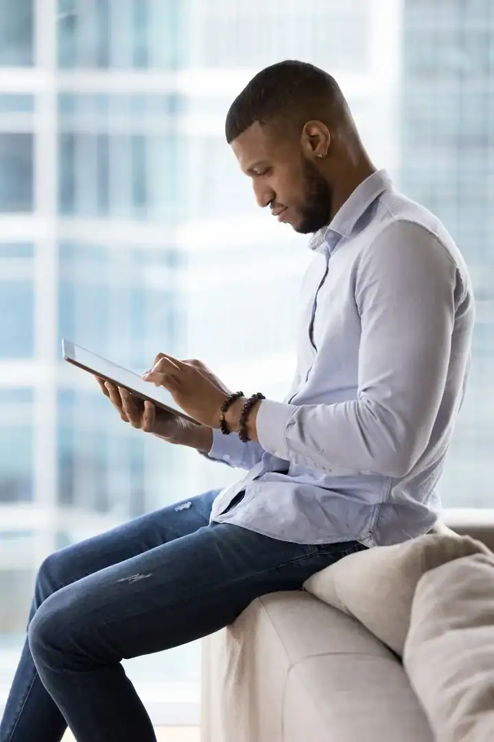 A man sits on the arm of a couch reading on a tablet, with a window behind him.
